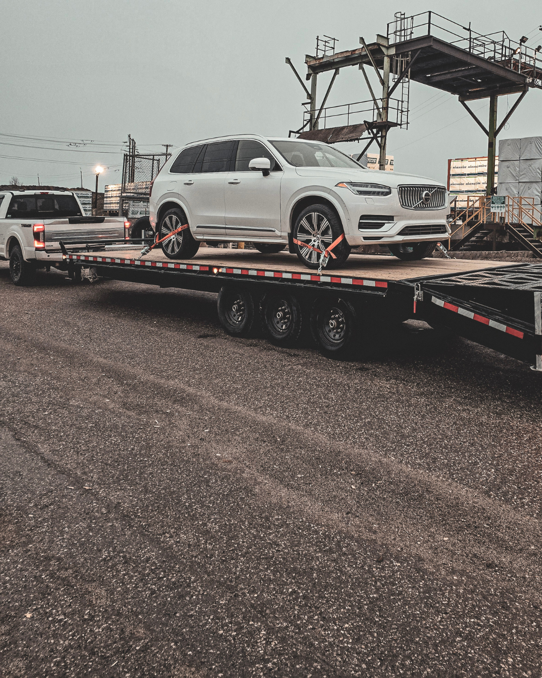 White Volvo XC90 luxury SUV securely strapped on a flatbed trailer, being towed by a white pickup truck in an industrial setting. The overcast sky and rugged surroundings add to the heavy-duty transportation theme. #CarTransport #LuxurySUV #VolvoXC90 #AutoHauling